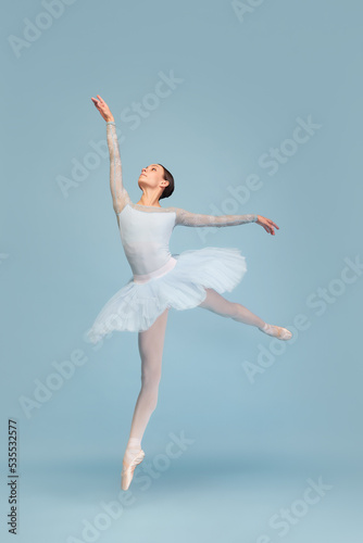 Portrait of tender young ballerina dancing, performing isolated over blue studio background. Lightness and grace