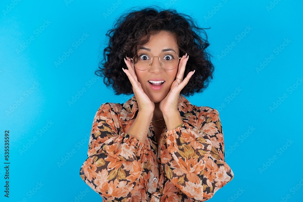 young brunette woman with curly hair wearing flowered dress standing over blue background Pleasant looking cheerful, Happy reaction young brunette woman with curly hair wearing flowered dress standing over blue background Pleasant looking cheerful, Happy reaction