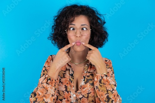 young brunette woman with curly hair wearing flowered dress standing over blue background crosses eyes and makes fish lips funny grimace photo