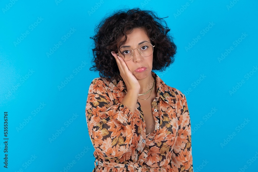 Sad lonely young brunette woman with curly hair wearing flowered dress standing over blue background touches cheek with hand bites lower lip and gazes with displeasure. Bad emotions Sad lonely young brunette woman with curly hair wearing flowered dress standing over blue background touches cheek with hand bites lower lip and gazes with displeasure. Bad emotions