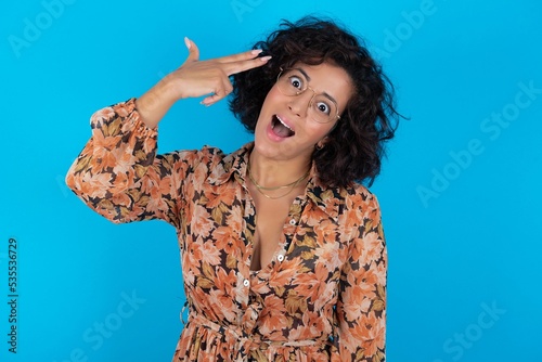 Obraz na plátně young brunette woman with curly hair wearing flowered dress standing over blue background foolishness around shoots in temple with fingers makes suicide gesture