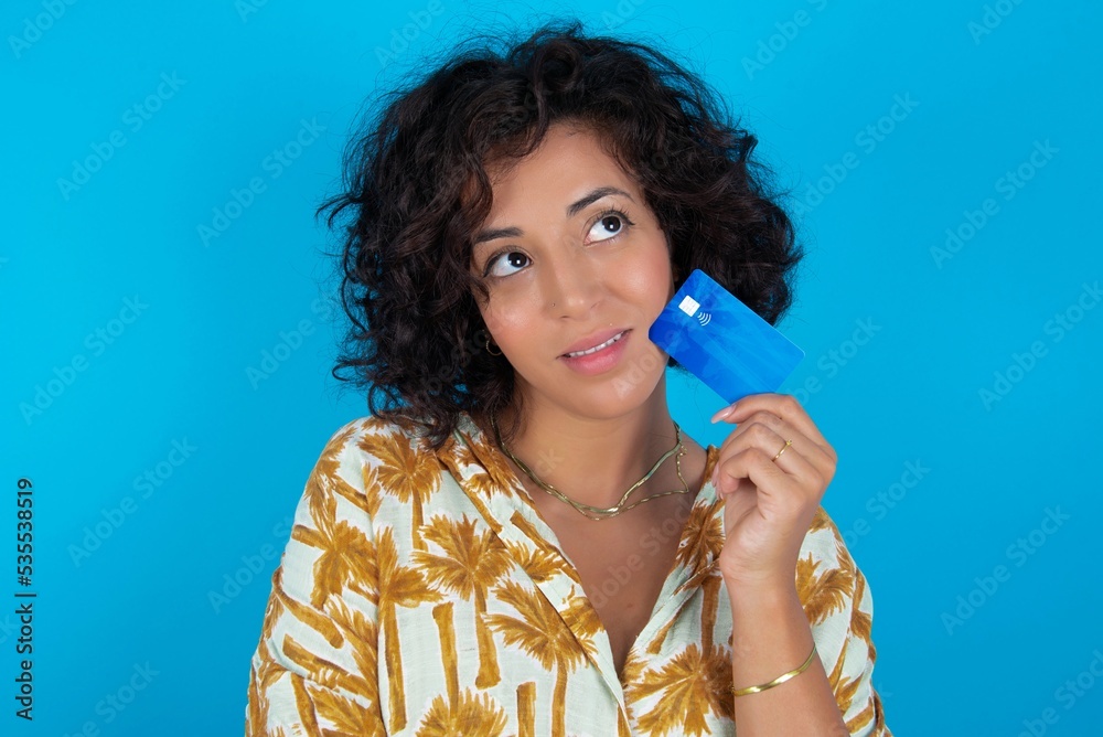 Lovely young brunette woman with curly hair wearing Hawaiian printed shirt standing over blue background showing credit card and looking away at copy space. Pensive Lovely young brunette woman with curly hair wearing Hawaiian printed shirt standing over blue background showing credit card and looking away at copy space. Pensive