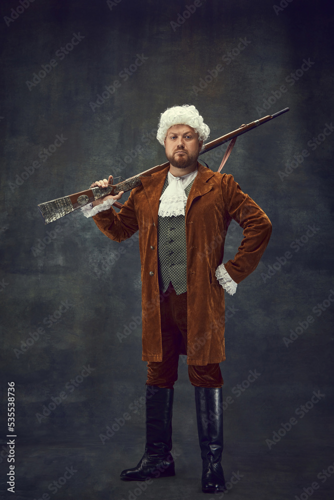 Fototapeta premium Creative portrait of retro style hunter in vintage hunting clothing with old gun isolated over dark background. Art, fashion, emotions, traditions, hobby concept