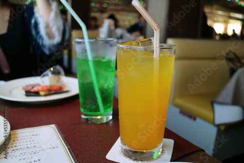 There are two glasses of brightly colored drinks on the table. Green Hami fruit juice and yellow orange juice are plugged into straws. Dining scene