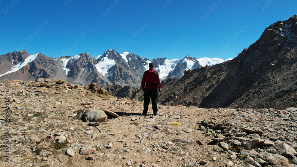 The guy looks at the mountains and snow-capped peaks. High mountains stretch to the blue sky. The peaks of the mountains are covered with glaciers. Steep cliffs down. There are large stones and moss
