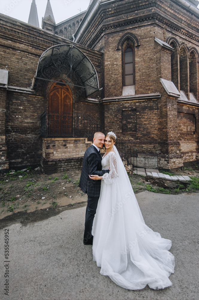 Obraz premium A young groom in a blue plaid suit and a cute bride in a white long dress with a diadem, a bouquet, stand embracing, near the church, the temple against the sky. wedding photography.