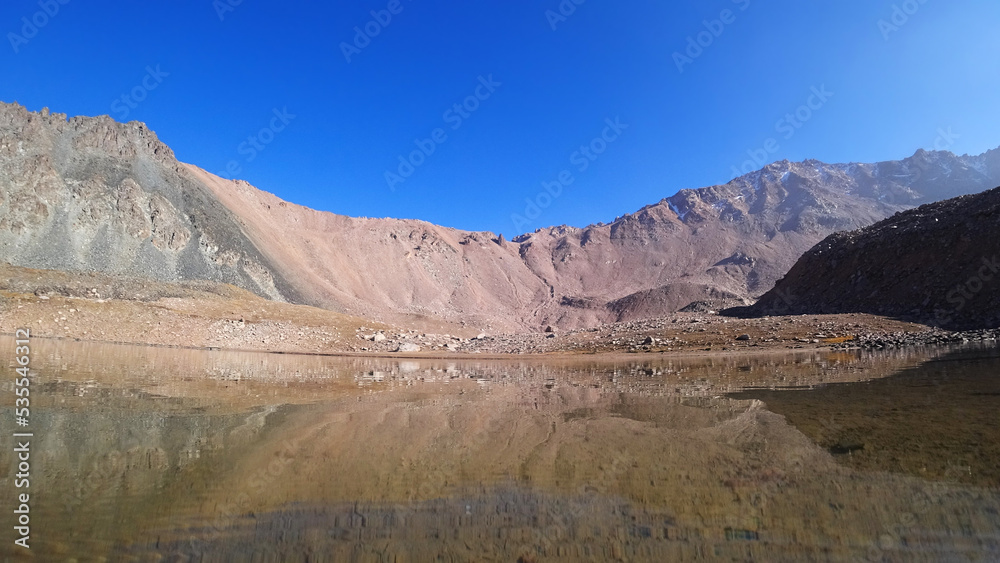 Transparent mountain lake surrounded by rocks. The water is like a mirror, reflecting the rocks and the slope. Light ripples on the surface of the water. The bottom is visible. There are big stones