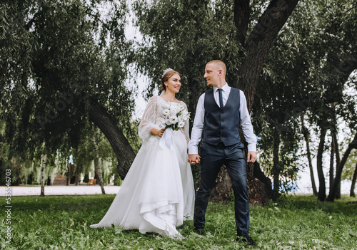 A stylish groom in a blue vest and a beautiful smiling bride are walking in the park on the green grass, holding hands. Wedding photo of newlyweds in love.