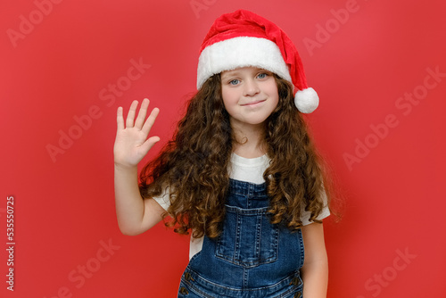 Portrait of beautiful smiling little girl in Santa hat waving hand  positive looking at camera  posing isolated over red color background wall in studio. Happy New Year 2023 holiday and Xmas concept