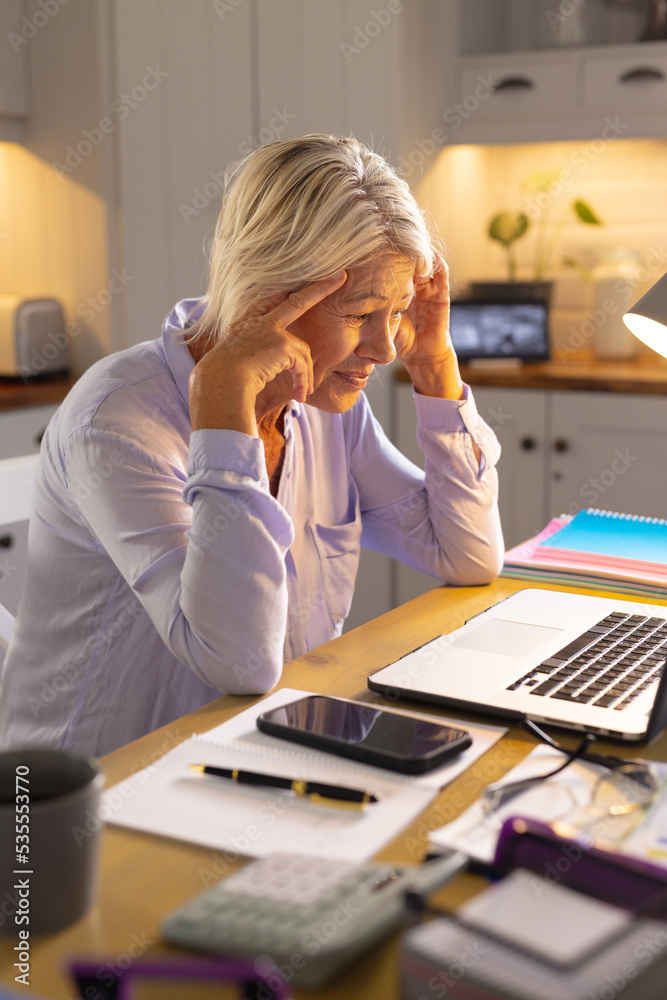 Thoughtful senior caucasian woman sitting at table in kitchen, using laptop Thoughtful senior caucasian woman sitting at table in kitchen, using laptop