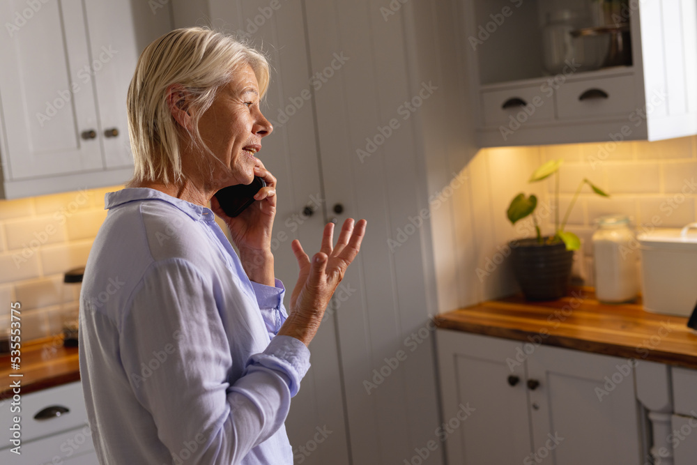 Happy senior caucasian woman standing in kitchen, talking on smartphone Happy senior caucasian woman standing in kitchen, talking on smartphone
