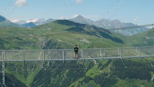 Man tourist outdoor traveler walking on a suspension metal bridge, taking photos on a bright summer day with landscape nature panoramic green mountains view high in the air, at high altitude in Alps. (ID: 535553729)