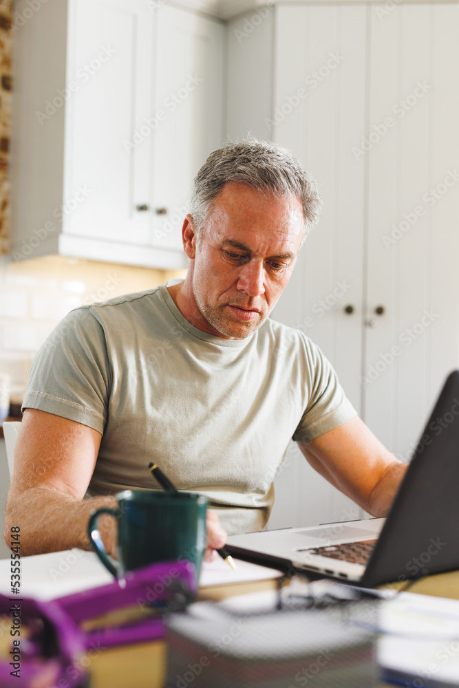 Happy caucasian man sitting at table in kitchen and using laptop and taking notes Happy caucasian man sitting at table in kitchen and using laptop and taking notes
