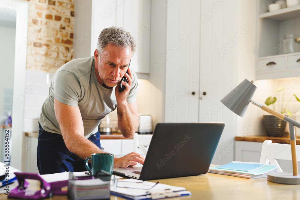 Happy caucasian man sitting at table in kitchen and using laptop and talking on smartphone Happy caucasian man sitting at table in kitchen and using laptop and talking on smartphone