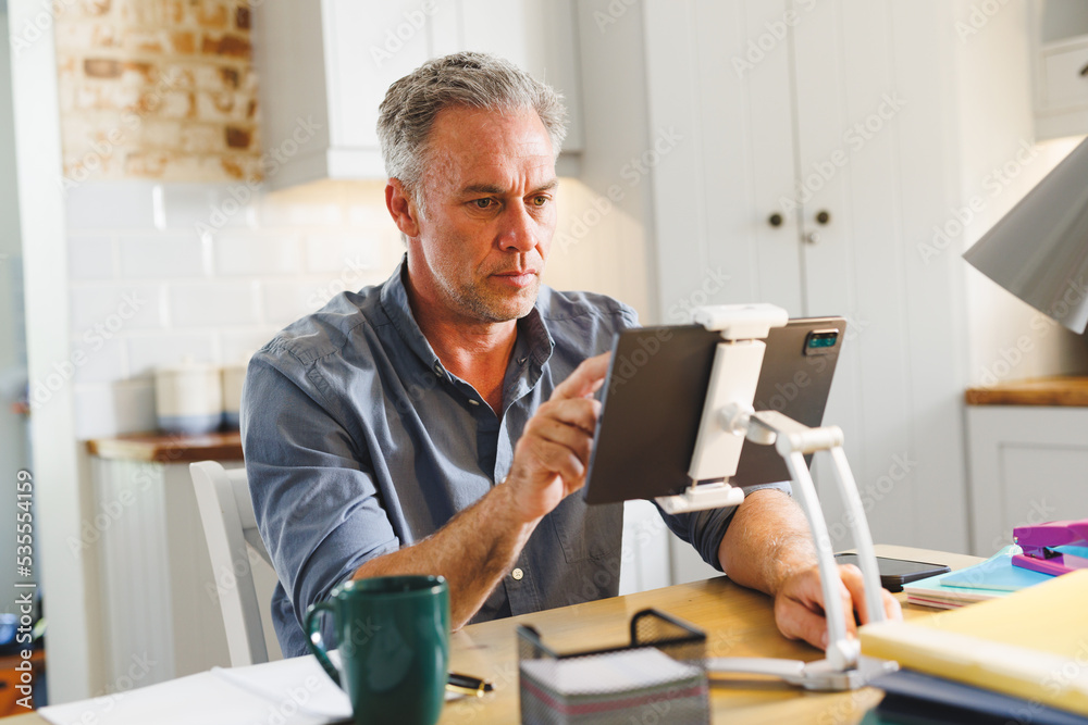 Happy caucasian man sitting at table in kitchen and using tablet