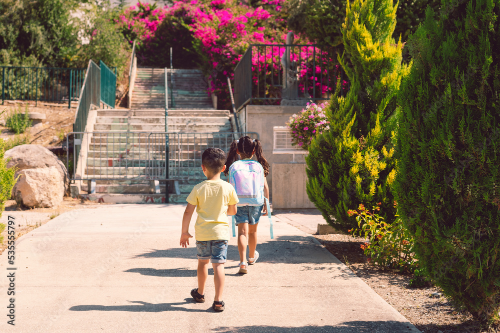 Fototapeta premium A young children going back to school. Little girl with a tie dye backpack walks to preschool with her brother at a sunny day.