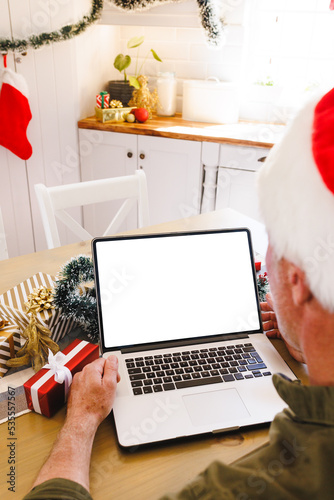 Caucasian man wearing santa hat  sitting at table in kitchen  using laptop with copyspace
