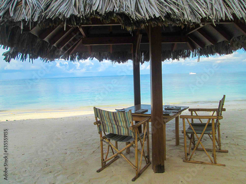 Fototapeta Naklejka Na Ścianę i Meble -  POV from a Tropical resort with no people on the beach and sun umbrella over a table with chairs,  horizon over water in a cloudy sky