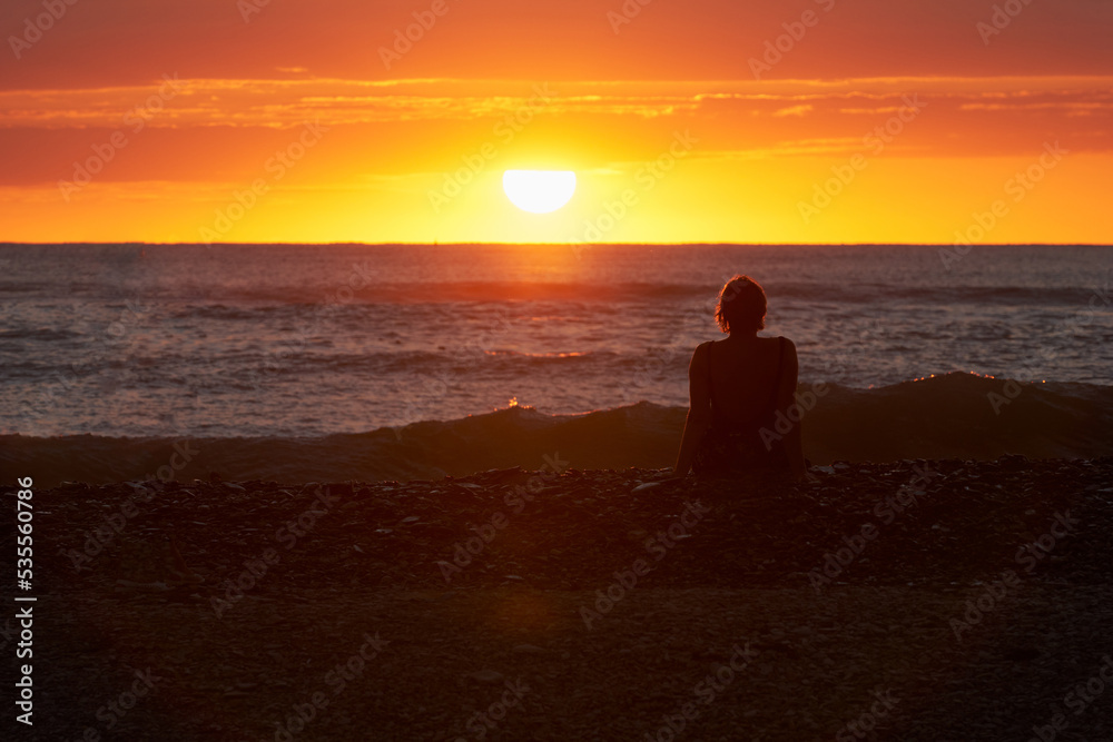 A picturesque poppy sunset over the sea. A woman is sitting on the shore and admiring the setting sun. Selective focus.