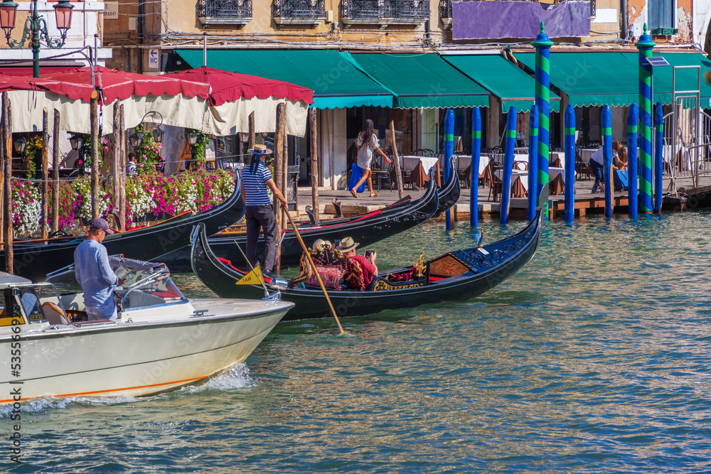 A gondolier standing in a gondola steers a boat with an oar in his hands, an elderly couple of tourists in a gondola filming a journey on smartphones along the coast of a Venice city street