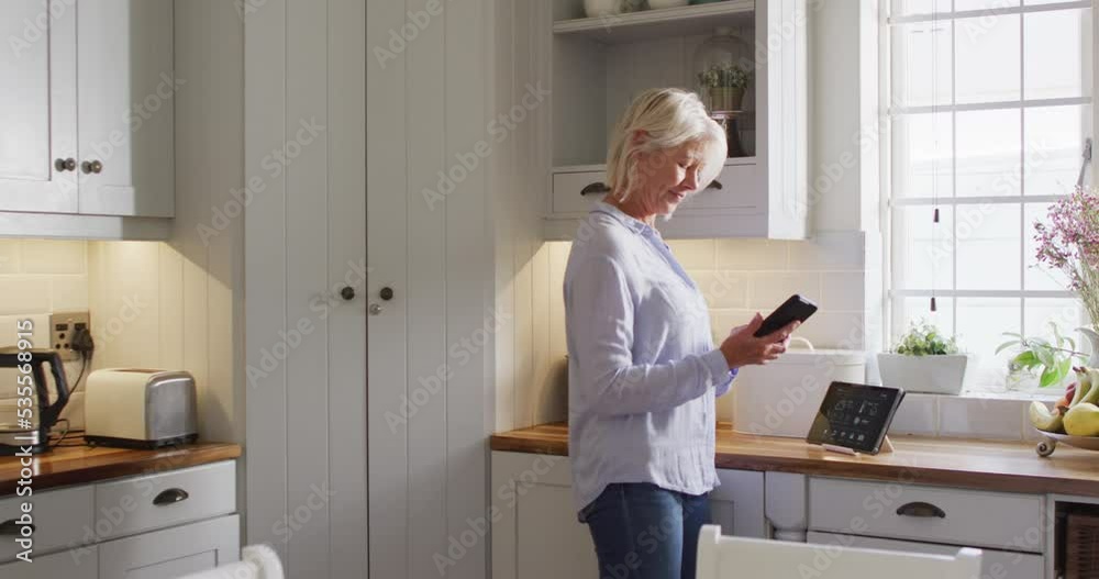 Happy senior caucasian woman standing in kitchen, using smartphone and tablet Happy senior caucasian woman standing in kitchen, using smartphone and tablet