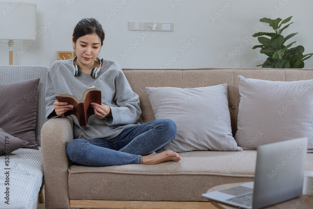 Happy Asian young woman wearing headphones with a book sitting relaxing on sofa at home