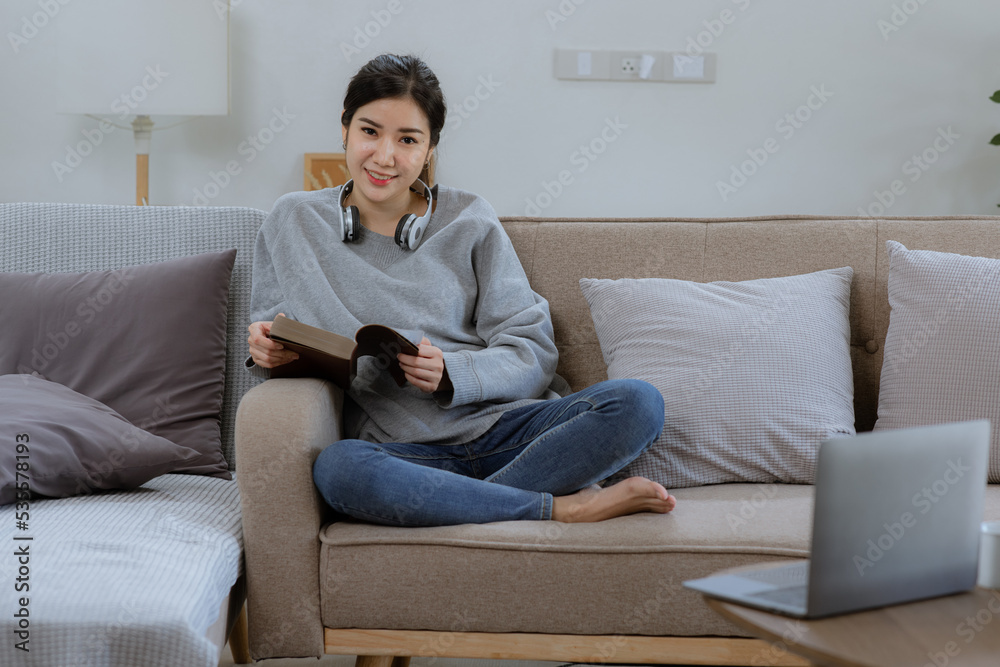 Happy Asian young woman wearing headphones with a book sitting relaxing on sofa at home Happy Asian young woman wearing headphones with a book sitting relaxing on sofa at home