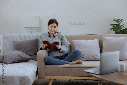 Happy Asian young woman wearing headphones with a book sitting relaxing on sofa at home