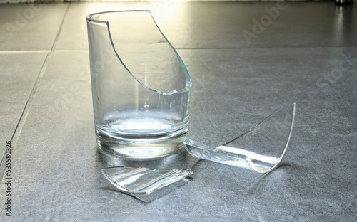 Closeup of empty cracked drinking glass isolated on grey tile background with sunlight, beverage glass cruash small shattered piece. Accident in kitchen and shard of mirror will be danger