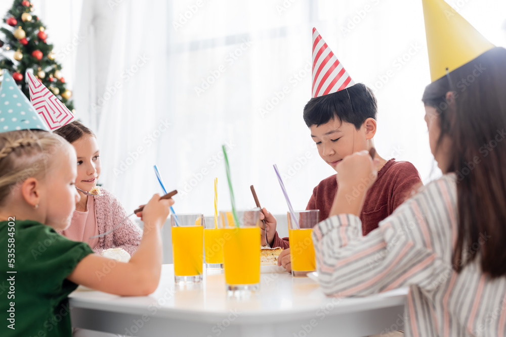 Asian boy in party cap looking at birthday cake near friends and orange juice in living room Asian boy in party cap looking at birthday cake near friends and orange juice in living room