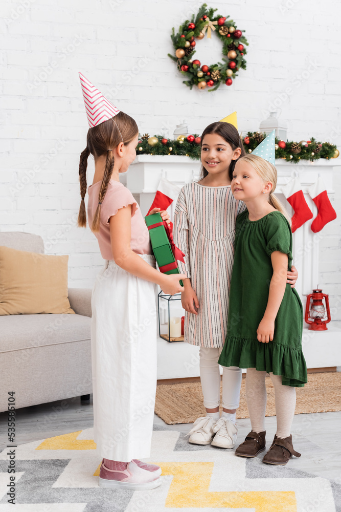 Obraz premium Girls in party caps looking at friend with gift box near blurred christmas stockings at home