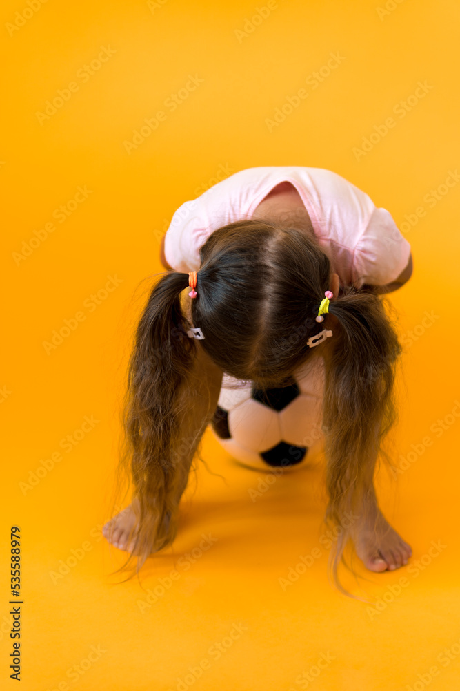 Fototapeta premium Authentic cute smiling preschool little girl with classic black and white soccer ball look at camera on yellow background. child play football in t-shirt and shorts. Sport, championship, team concept