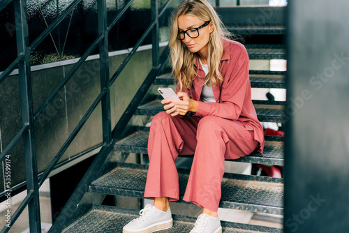 Young casual woman using smartphone while sitting on the iron steps of a modern office building. photo