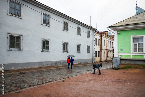 Couple of people under umbrella walk along the street of small old town among retro houses, signboard in form of bull 