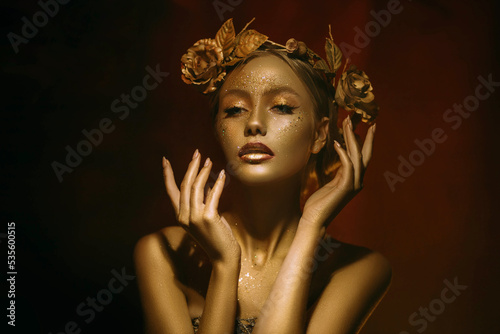 Portrait Closeup Beauty fantasy woman, face in gold paint. Golden shiny skin. Fashion model girl, image goddess. Glamorous crown, wreath roses, jewellery accessories. Professional metallic makeup