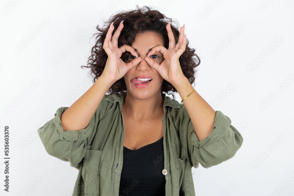 young beautiful woman with curly short hair wearing green overshirt over white wall doing ok gesture like binoculars sticking tongue out, eyes looking through fingers. Crazy expression. young beautiful woman with curly short hair wearing green overshirt over white wall doing ok gesture like binoculars sticking tongue out, eyes looking through fingers. Crazy expression.