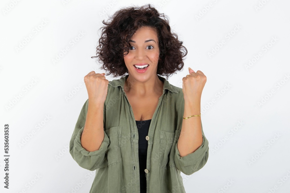 Obraz na plátně young beautiful woman with curly short hair wearing green overshirt over white wall celebrating surprised and amazed for success with arms raised and open eyes