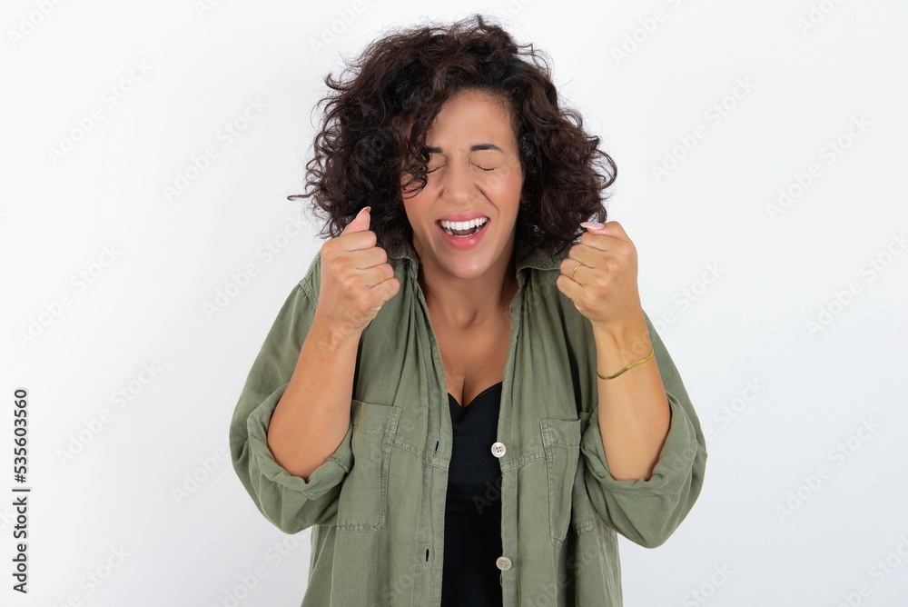 Φωτογραφία young beautiful woman with curly short hair wearing green overshirt over white wall being excited for success with raised arms and closed eyes celebrating victory