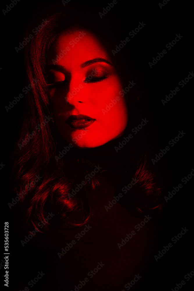 Beauty and fashion concept. Beautiful woman portrait standing in darkness with illuminated face. Model with evening make-up with closed eyes and wavy hair. Toned image with red color