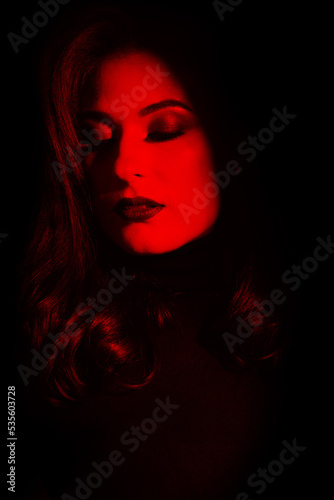 Beauty and fashion concept. Beautiful woman portrait standing in darkness with illuminated face. Model with evening make-up with closed eyes and wavy hair. Toned image with red color