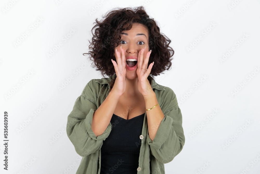 young beautiful woman with curly short hair wearing green overshirt over white wall shouting excited to front. young beautiful woman with curly short hair wearing green overshirt over white wall shouting excited to front.