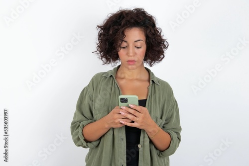 Excited young beautiful woman with curly short hair wearing green overshirt over white wall  winking and eye hold smart phone use read social network news