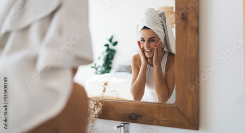 Young beautiful female with towel on head smiling to mirror reflection standing in the bathroom at home. 30 years old happy woman doing daily morning rituals and cleansing. Enjoying healthy skin care
