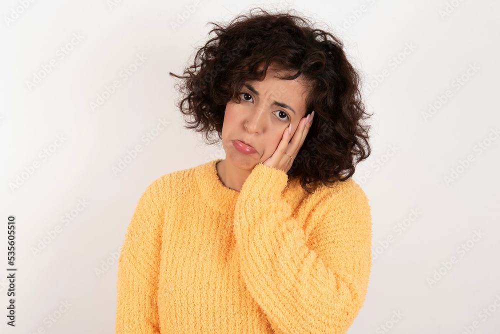 Sad lonely young beautiful woman with curly short hair wearing yellow sweater over white background touches cheek with hand bites lower lip and gazes with displeasure. Bad emotions