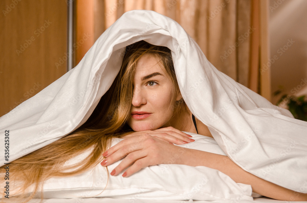 Beautiful young woman in morning in white bed in rays of sun, covered with blanket with her head, playfully smiling Beautiful young woman in morning in white bed in rays of sun, covered with blanket with her head, playfully smiling
