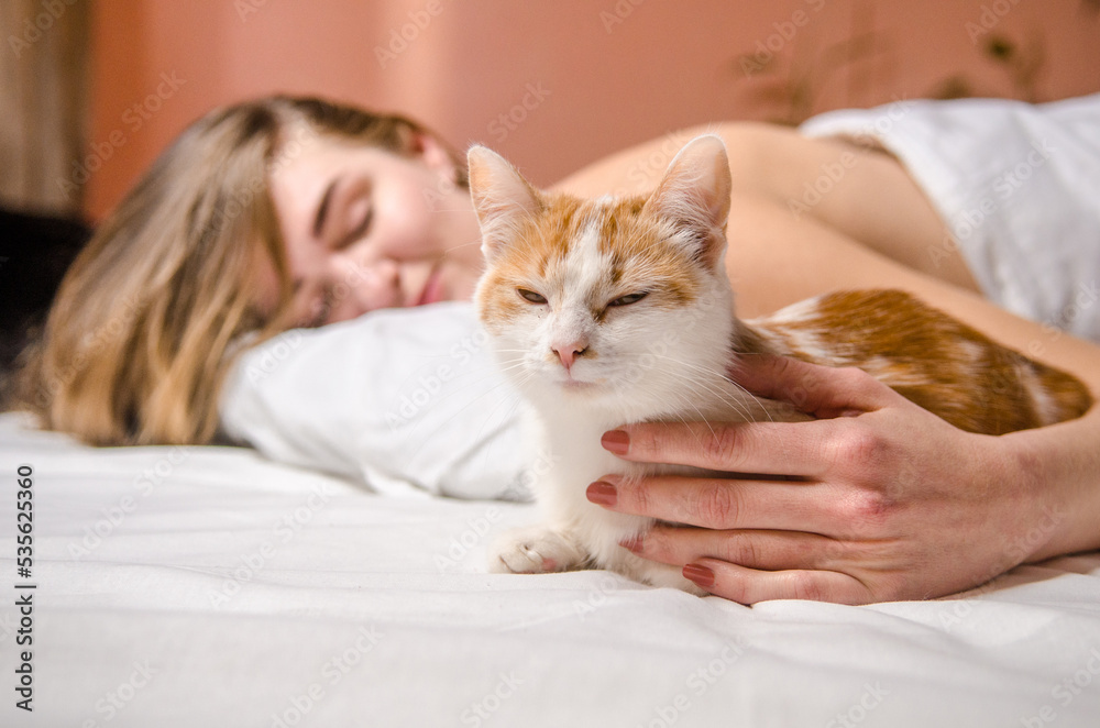Beautiful young woman lies in white bed with a ginger cat, smiles, kisses and hugs her, love animals, friendship