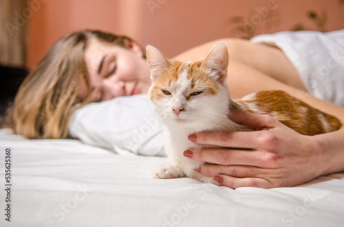 Beautiful young woman lies in white bed with a ginger cat, smiles, kisses and hugs her, love animals, friendship