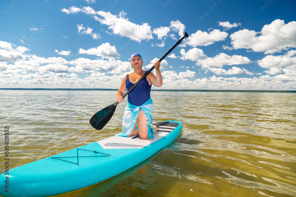 A woman in a pareo with a paddle on her knees on a SUP board in the lake against the background of white clouds on a clear blue sky.