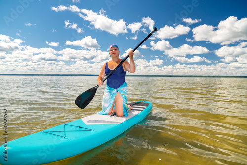 A woman in a pareo with a paddle on her knees on a SUP board in the lake against the background of white clouds on a clear blue sky.
