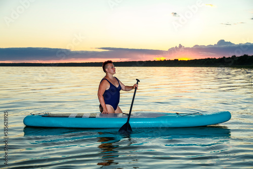 A woman in a swimsuit is kneeling on a SUP board with a paddle in the lake on a sunny day against the backdrop of white clouds.